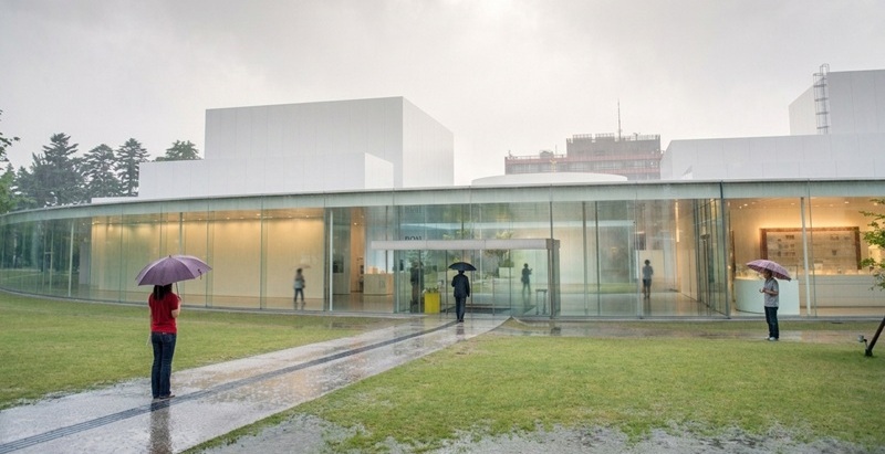 雨の日の金沢21世紀美術館。円形のガラス張り外観と濡れた芝生。傘をさした来館者が入口に向かう