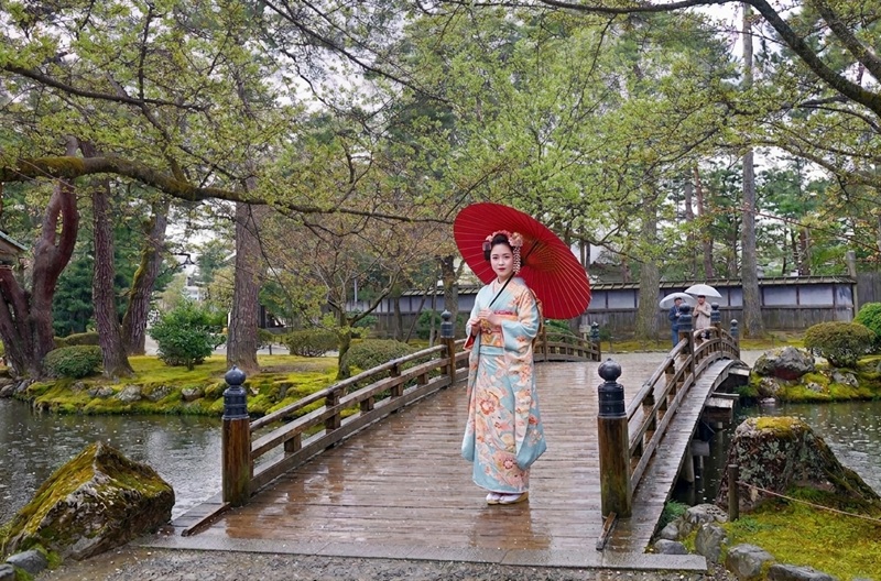 雨の金沢・兼六園の橋に立つ着物姿の女性。新緑と苔に覆われた庭園に赤い傘が映える雨の日の風情